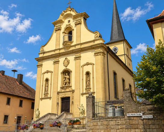 Sankt Matthäus (Rittershausen) Das Bild zeigt die Saankt-Matthäus-Kirche in Rittershausen mit barocker Fassade, einem spitzen Turm, mehreren Statuen und einem zentralen Eingang, umgeben von grünen Bäumen unter blauem Himmel.