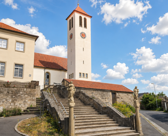 Sankt Bartholomäus (Acholshausen) Die Aufnahme zeigt die Kirche Sankt Bartholomäus in Acholshausen mit hohem Turm und Uhr, umgeben von historischen Gebäuden mit roten Ziegeldächern und blumengeschmückten Fenstern, sowie eine breite Steintreppe. Himmel ist blau mit Wolken.