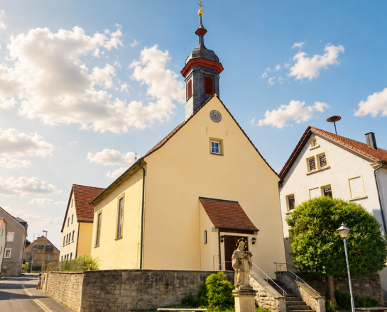 Sankt Peter und Paul (Sächsenheim) Das Bild zeigt die Peter-und-Paul-Kirche in Sächsenheim mit hohem Turm und Uhr, umgeben von traditionellen Häusern mit roten Ziegeldächern unter blauem Himmel mit weißen Wolken.