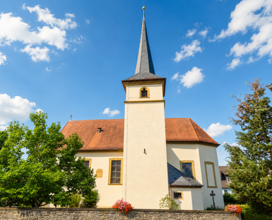Sankt Andreas (Bolzhausen) Auf dem Bild sieht man die Sankt-Andreas-Kirche in Bolzhausen mit spitzem Turm und rotem Dach, umgeben von Häusern und Bäumen unter blauem Himmel mit wenigen Wolken.