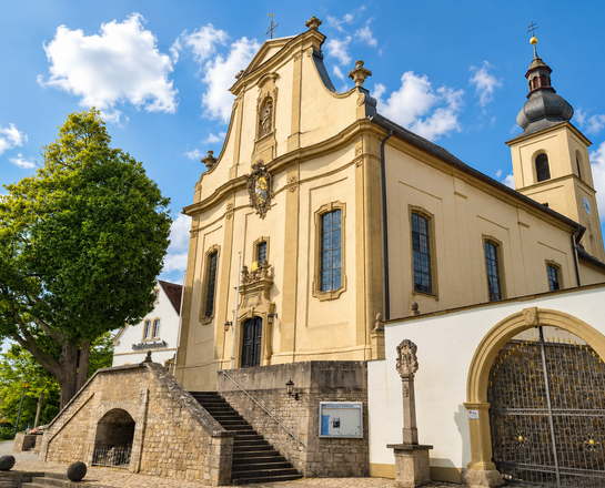 Sankt Johannes der Täufer (Sonderhofen) Das Bild zeigt die Sankt-Johannes-Kirche in Sonderhofen mit barocker Fassade, einem Turm mit dunkler Kuppel und einem verzierten Eingangsportal auf einem gepflasterten Platz.