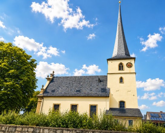 Sankt Markus und Mariä Verkündigung Das Bild zeigt die Sankt-Markus-Kirche in Wolkshausen mit hohem Turm, spitzem Dach und Uhr unter blauem Himmel.