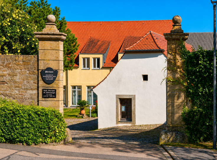 Pfarrbüro Gaukönigshofen Das Bild zeigt den Eingang zum Pfarrhof in Gaukönigshofen mit rotem Ziegeldach und gelblichen Wänden. Es hat ein Schild mit Öffnungszeiten und ist von Grünpflanzen umgeben.